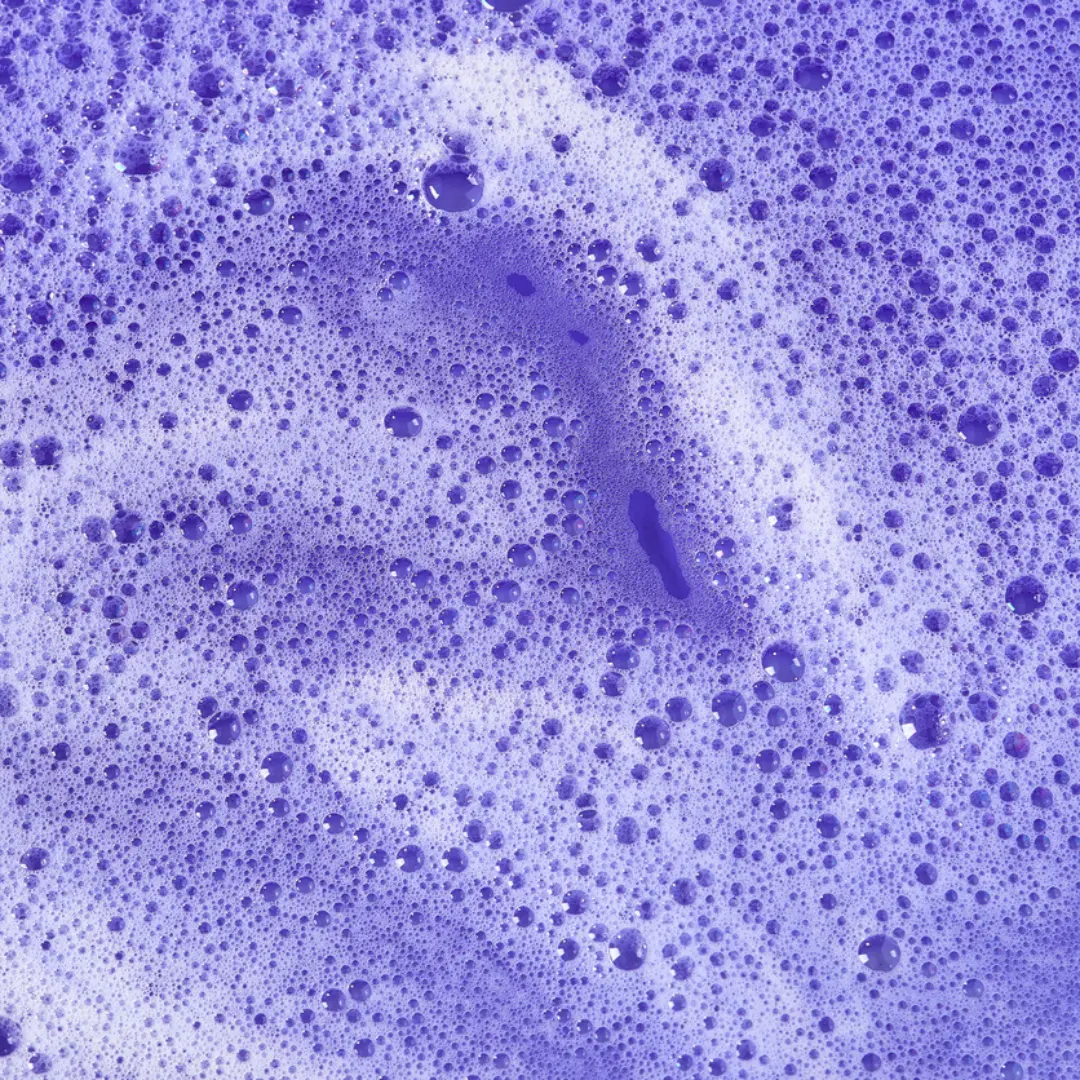 Des bulles de savon violettes et de la mousse recouvrent une surface, formant un motif abstrait texturé et pétillant, évoquant l'apparence du Shampoing Anti-Jaunissement S4, où la qualité et le prix rencontrent l'inspiration créative. Dahlia Color Professionnel