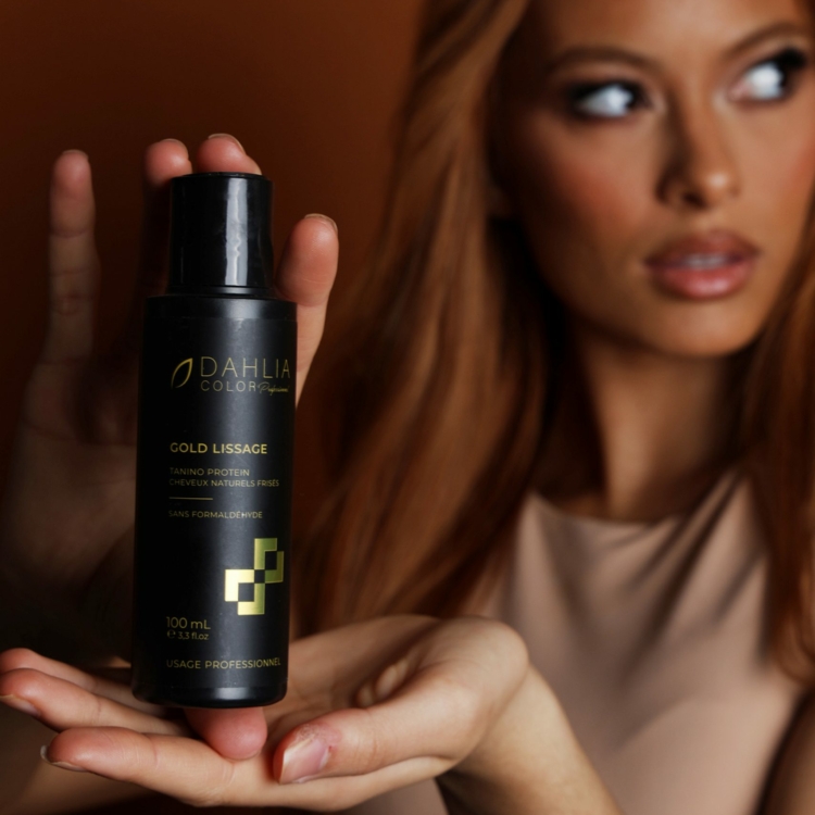 Femme présentant une bouteille de produit capillaire Dahlia Gold Lissage sur fond marron. Dahlia Color Professionnel