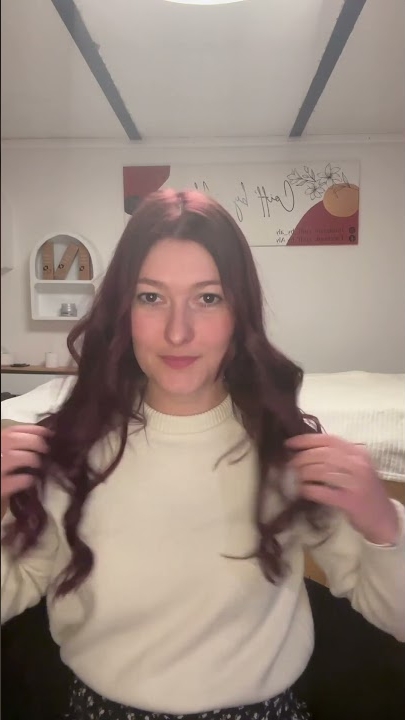 Une femme aux cheveux ondulés portant un pull blanc est assise à l'intérieur, regardant la caméra et souriant légèrement. Dahlia Color Professionnel