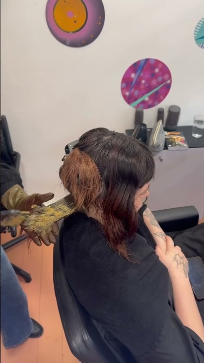 Un coiffeur applique de l'eau de Javel sur les cheveux d'une femme dans un salon, avec des outils de salon visibles en arrière-plan. Dahlia Color Professionnel