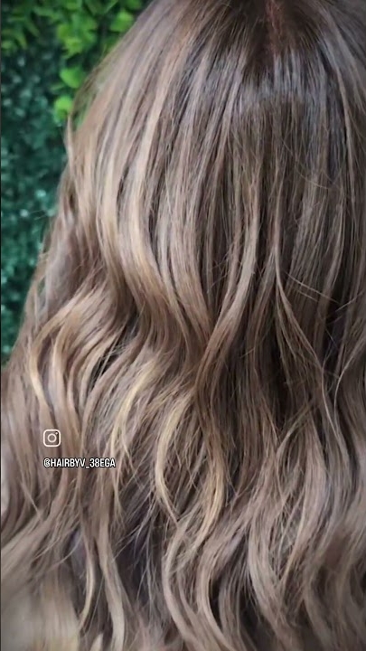 Cheveux ondulés châtain clair avec des reflets subtils devant un feuillage vert. Dahlia Color Professionnel